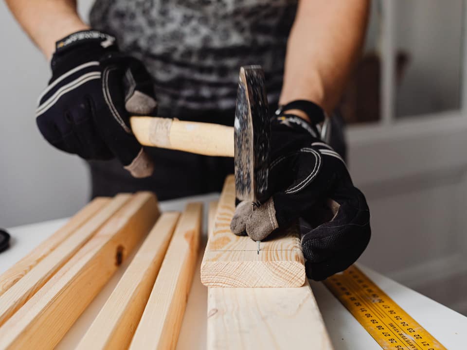 A gloved hand driving a nail into a length of framing timber.