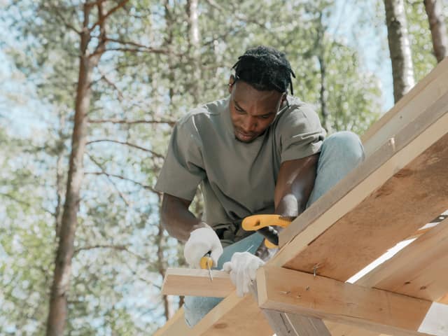 A carpenter fastening boards to a timber frame outdoors on a bright day.